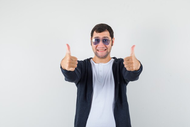 Jovem macho em t-shirt, jaqueta mostrando dois polegares para cima e olhando alegre, vista frontal.