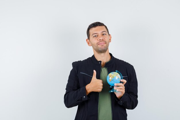 Jovem macho em t-shirt, jaqueta segurando o globo com o polegar para cima e parecendo satisfeito, vista frontal.