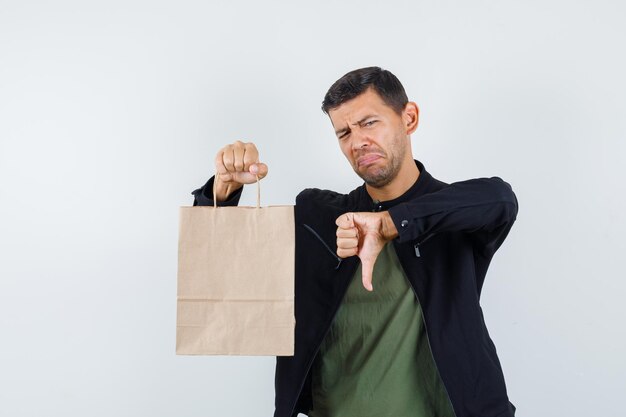 Jovem macho em t-shirt, jaqueta segurando o saco de papel com o polegar para baixo e olhando descontente, vista frontal.