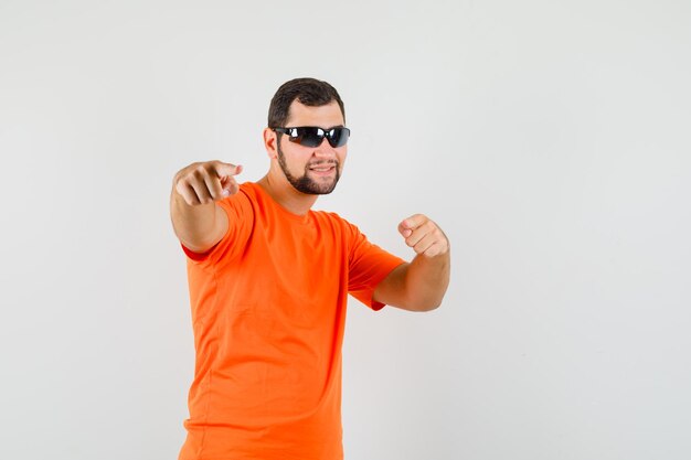 Jovem macho em t-shirt laranja, apontando para a câmera e olhando bonito, vista frontal.