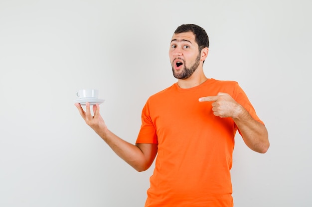 Jovem macho em t-shirt laranja apontando para a xícara com Pires e olhando a vista frontal positiva.