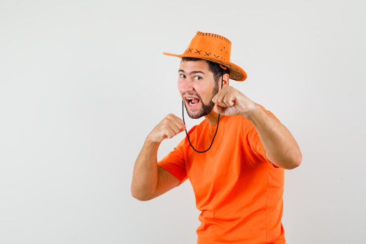 Foto grátis jovem macho em t-shirt laranja, chapéu em pé em pose de luta e olhando poderoso, vista frontal.