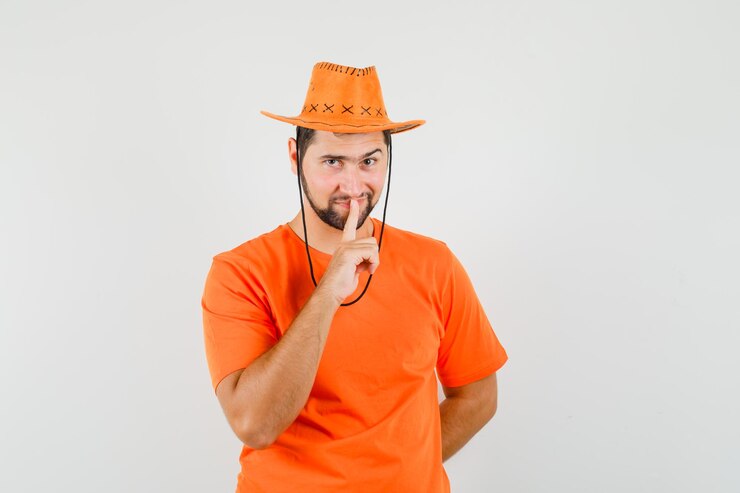 Foto grátis jovem macho em t-shirt laranja, chapéu mostrando gesto de silêncio e olhando astuto, vista frontal.