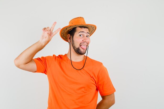 Jovem macho em t-shirt laranja, chapéu mostrando sinal de vitória, mostrando a língua e olhando engraçado, vista frontal.