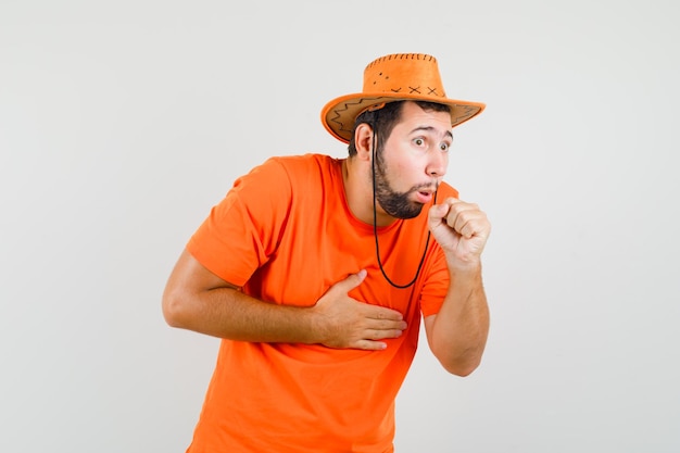 Jovem macho em t-shirt laranja, chapéu, sofrendo de tosse e parecendo doente, vista frontal.