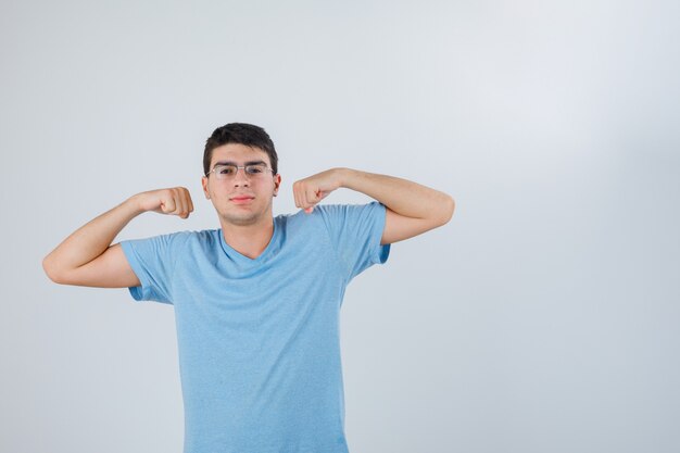 Jovem macho em t-shirt mostrando gesto de músculos e olhando confiante, vista frontal.