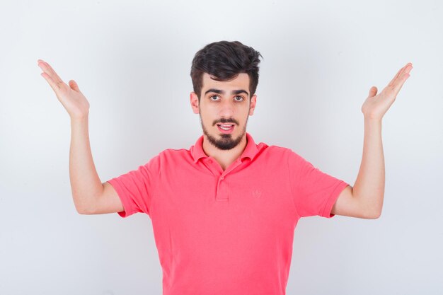 Jovem macho em t-shirt rosa, levantando as mãos e olhando confiante, vista frontal.