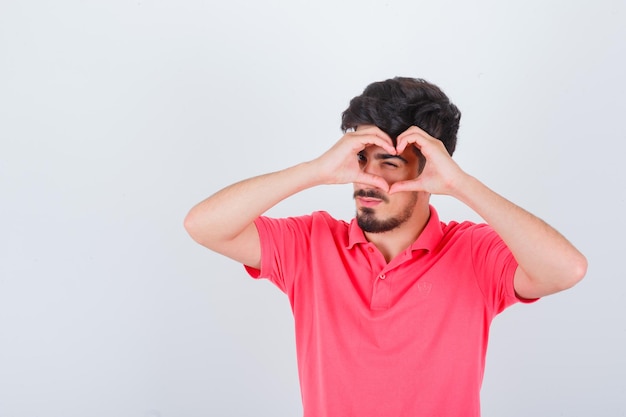 Jovem macho em t-shirt rosa mostrando um gesto de coração e olhando confiante, vista frontal.