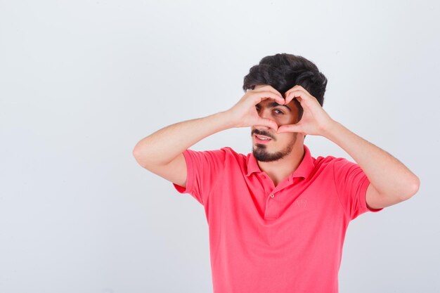 Jovem macho em t-shirt rosa mostrando um gesto de coração e olhando confiante, vista frontal.