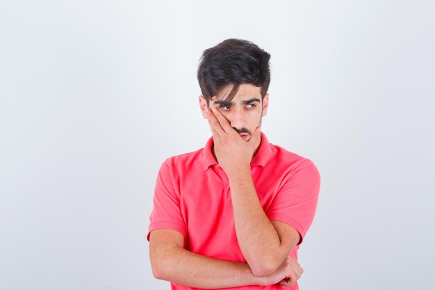 Jovem macho em t-shirt, segurando a mão no rosto e olhando hesitante, vista frontal.