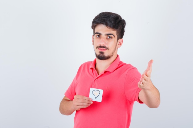 Jovem macho em t-shirt segurando nota enquanto levanta a mão de forma questionadora e parece confiante, vista frontal.