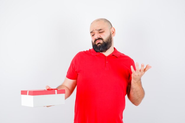 Jovem macho em t-shirt vermelha, esticando a mão em um gesto de questionamento e parecendo insatisfeito, vista frontal.