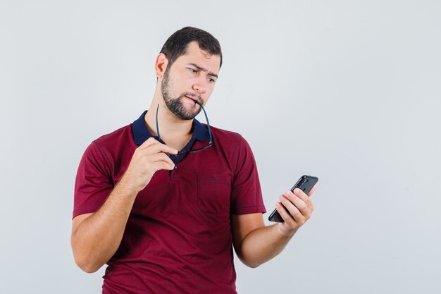 Jovem macho em t-shirt vermelha, olhando para o telefone e olhando perplexo, vista frontal.