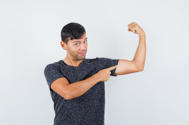 Jovem macho em uma camiseta preta, apontando para os músculos do braço e parecendo confiante, vista frontal.