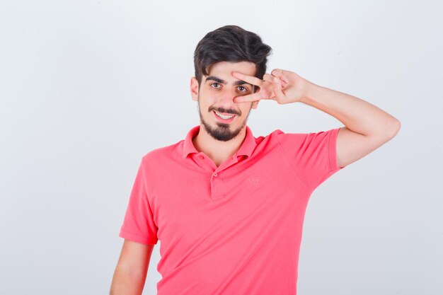 Jovem macho em uma camiseta rosa mostrando o sinal de V no olho e olhando alegre, vista frontal.