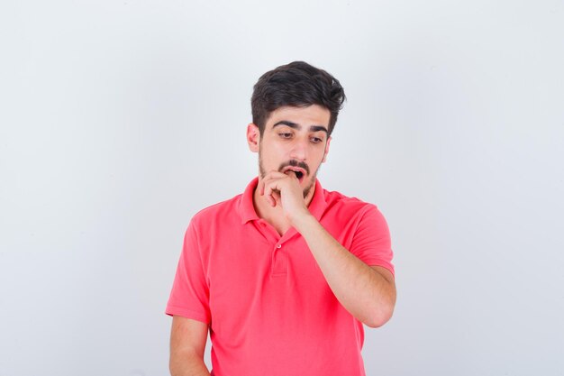 Jovem macho em uma camiseta rosa, segurando o dedo na boca e parecendo desconfortável, vista frontal.