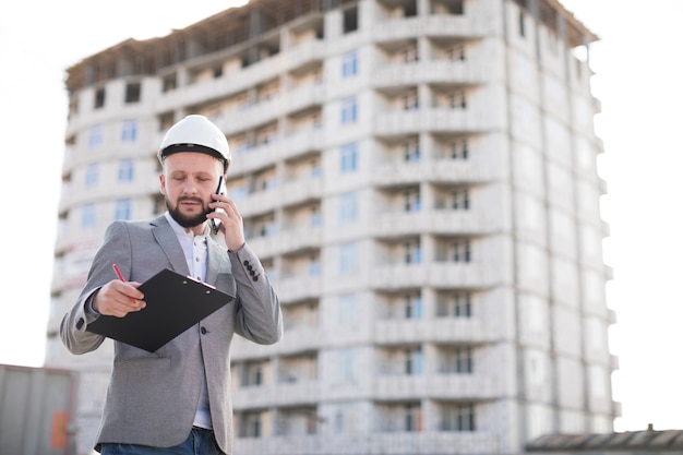 Jovem, macho, engenheiro, falando, ligado, cellphone, enquanto, segurando clipboard, em, local construção