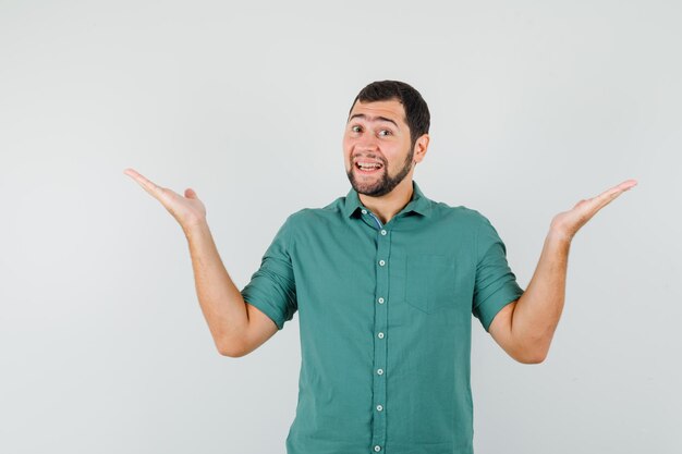 Jovem macho espalhando as palmas das mãos como segurando ou mostrando algo na camisa verde e olhando feliz, vista frontal.