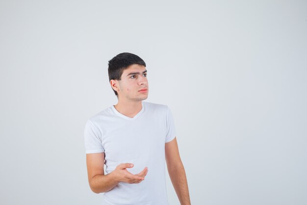 Jovem macho, esticando a mão em um gesto de questionamento em t-shirt e olhando pensativo, vista frontal.