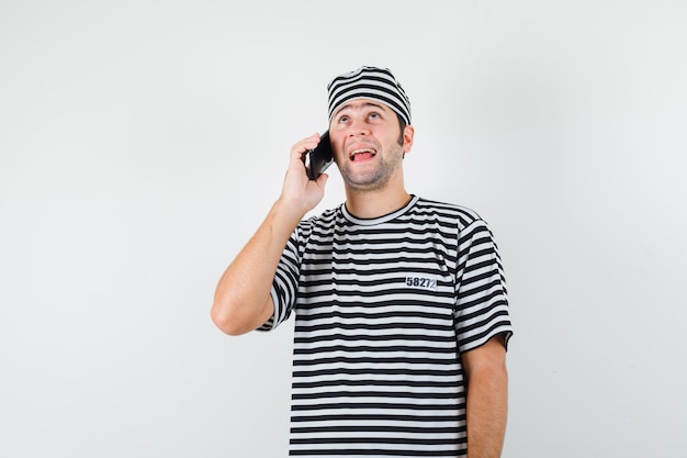 Jovem macho falando no celular em t-shirt, chapéu e olhando alegre, vista frontal.