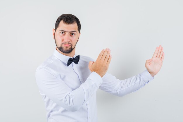 Jovem macho fazendo gesto de kung fu em camisa branca e parecendo com raiva. vista frontal.