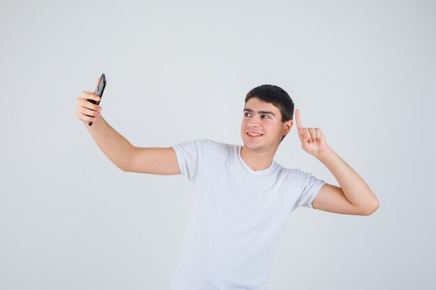 Jovem macho fazendo selfie enquanto aponta para cima em t-shirt e olhando alegre, vista frontal.