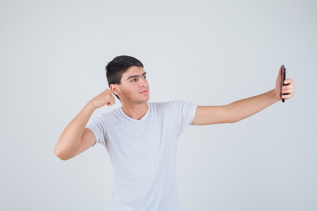 Jovem macho fazendo selfie enquanto mostra os músculos dos braços em uma camiseta e parece alegre. vista frontal.