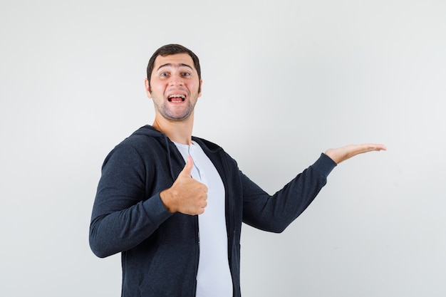 Jovem macho fingindo segurar algo, aparecendo o polegar em uma camiseta, jaqueta e parecendo alegre. vista frontal.