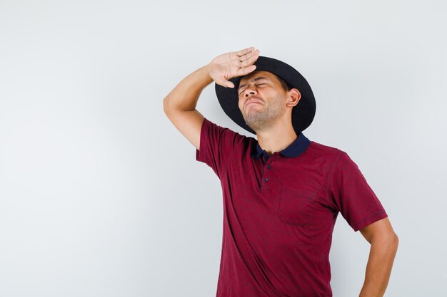 Jovem macho levantando a mão para evitar o sol forte na t-shirt, chapéu e parecendo tonto, vista frontal.