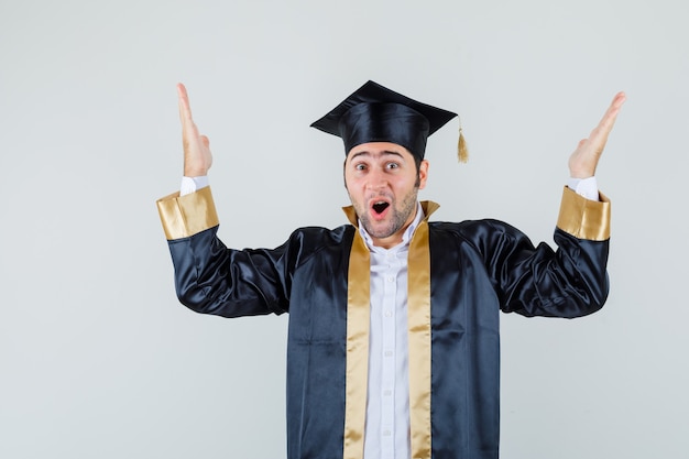 Jovem macho levantando as mãos em uniforme de pós-graduação e olhando feliz, vista frontal.