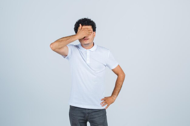 Jovem macho mantendo a mão nos olhos em camiseta branca, calça e parecendo calmo. vista frontal.