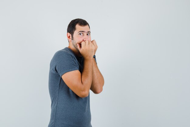 Jovem macho mordendo os punhos emocionalmente em uma camiseta cinza e parecendo assustado