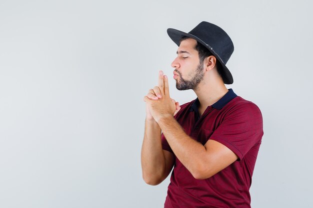 Jovem macho mostrando a pistola assinar em t-shirt, chapéu e olhando confiante, vista frontal.