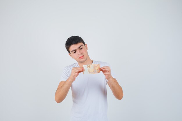 Jovem macho mostrando eurobanknote em t-shirt e olhando cuidadoso, vista frontal.