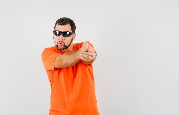 Jovem macho mostrando gesto de arma apontado em t-shirt laranja e olhando confiante, vista frontal.