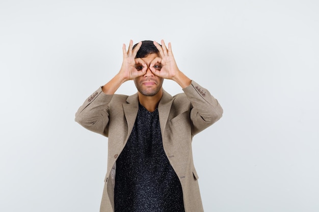 Jovem macho mostrando gesto de óculos com os dedos na jaqueta marrom acinzentada, camisa preta e parecendo engraçado. vista frontal.