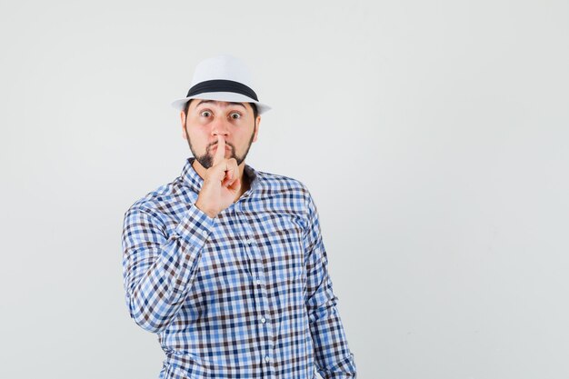 Jovem macho mostrando gesto de silêncio em camisa xadrez, chapéu e parecendo preocupado. vista frontal.