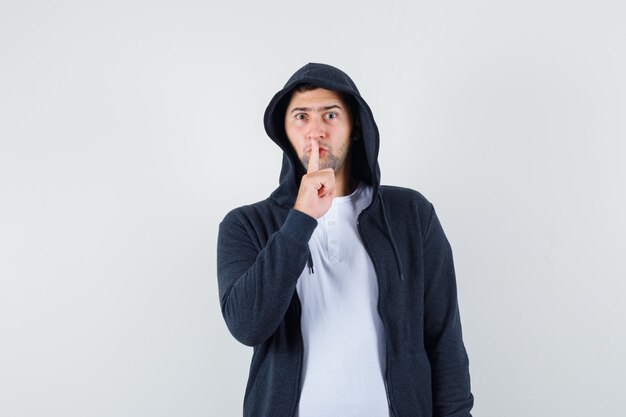 Jovem macho mostrando gesto de silêncio em t-shirt, jaqueta e olhando sério. vista frontal.