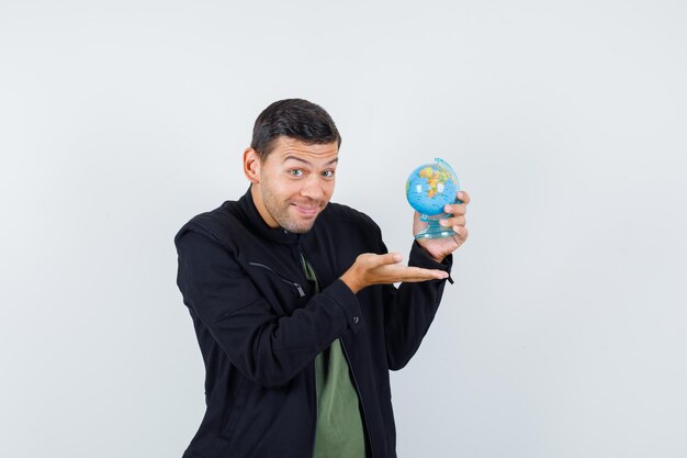 Jovem macho mostrando o globo do mundo em t-shirt, jaqueta e olhando alegre, vista frontal.