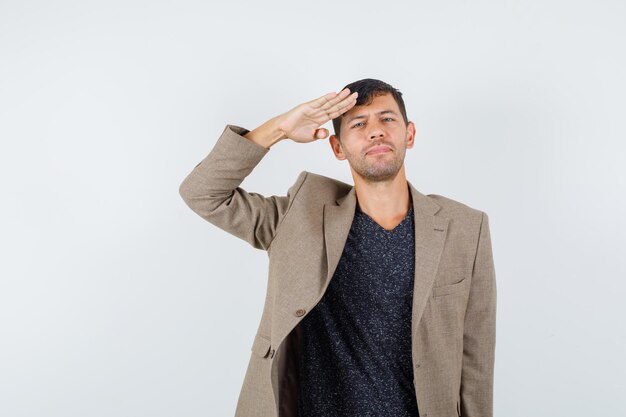 Jovem macho mostrando um gesto de Olá militar na jaqueta marrom acinzentada e olhando sério, vista frontal.