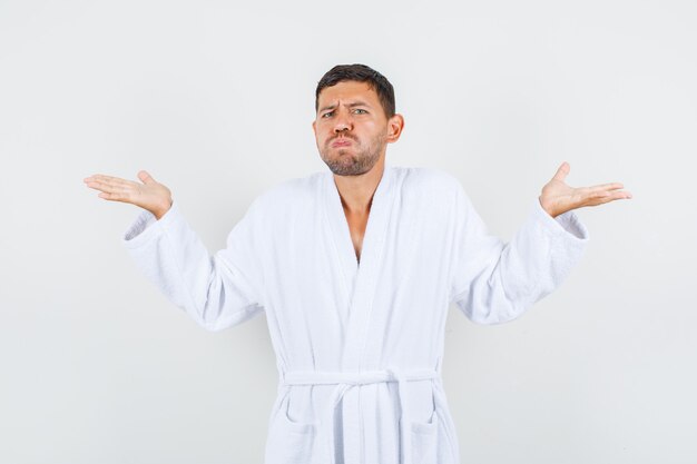 Jovem macho mostrando um gesto impotente em um roupão branco e parecendo confuso, vista frontal.