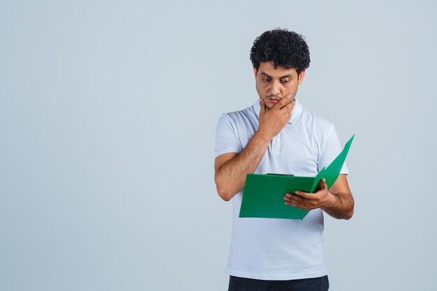 Jovem macho olhando as notas na prancheta em t-shirt branca, calça e olhando pensativo, vista frontal.