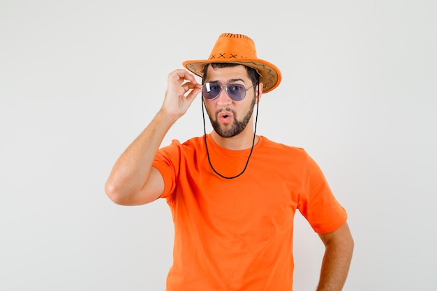 Jovem macho olhando através de óculos em t-shirt laranja, chapéu e parece legal. vista frontal.