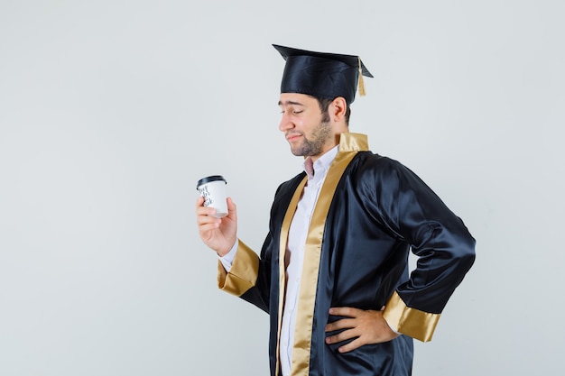 Jovem macho olhando para a xícara de café em uniforme de pós-graduação e olhando pensativo, vista frontal.