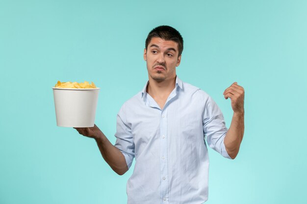 Jovem macho segurando a cesta com batata frita na superfície azul claro de frente