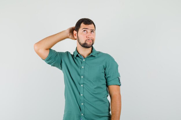 Jovem macho segurando a mão na cabeça enquanto olha para cima em uma camisa verde e parece pensativo. vista frontal.