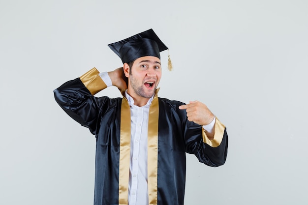 Jovem macho segurando a mão no pescoço, apontando para si mesmo em vista frontal do uniforme de pós-graduação.