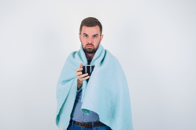 Foto grátis jovem macho segurando a taça enquanto olha para a câmera em camisa, jeans, cobertor e olhando sério. vista frontal.