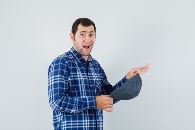 Jovem macho segurando boné na camisa azul e parecendo satisfeito. vista frontal.