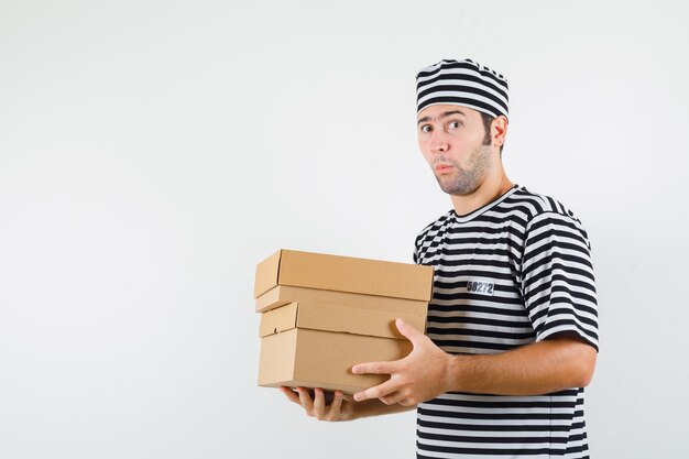 Jovem macho segurando caixas de papelão em t-shirt, chapéu e parecendo surpreso. vista frontal.
